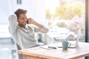 Freelance sorridente al telefono mentre lavora al computer da casa, con tazza e quaderno sul tavolo.