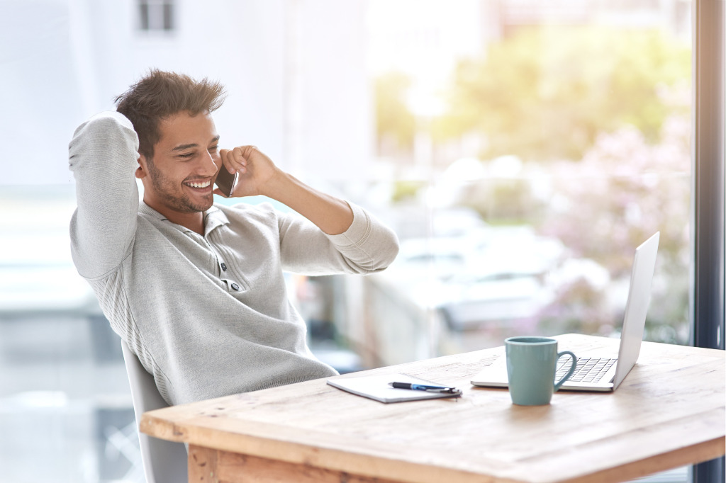 Freelance sorridente al telefono mentre lavora al computer da casa, con tazza e quaderno sul tavolo.