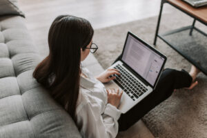 Donna che lavora da casa con un laptop, seduta a terra accanto al divano, simbolo del lavoro agile e dello smart working.
